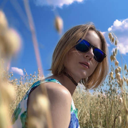 young attractive blonde woman in sunglasses sitting in oat field on sunny dayの写真素材