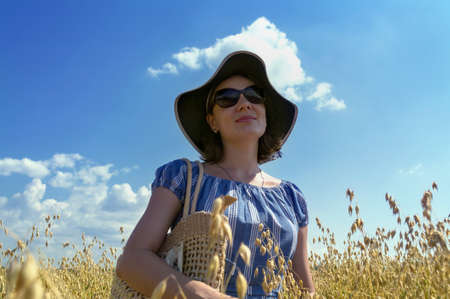 young brunette woman in hat with straw bag walking in oat filed view from belowの写真素材