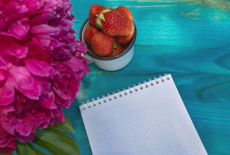 close up view of notepad and mug of strawberries on blue wooden table with part of peony bouquetの写真素材