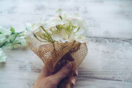 woman making small bouquet of hydrangea flowers decoratin it with golden netting on white wooden...