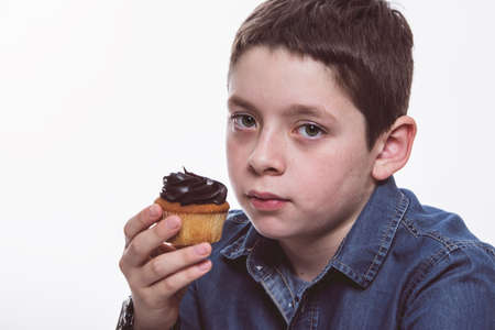 Young boy with denim shirt eating chocolate cupcake, on white isolated background. Close up...