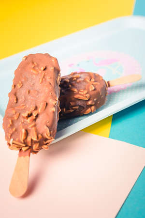 Dessert chocolate ice cream bars with nuts, wooden stick on blue and yellow pastel background....