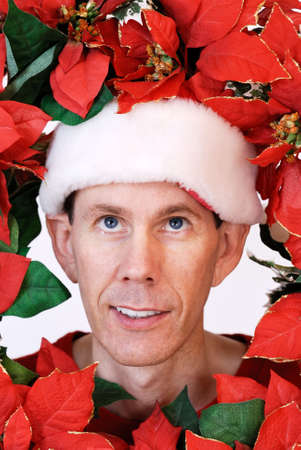 Man wearing Santa hat looking through the center of a poinsettia holiday wreath with his blue eyes glancing upward.の写真素材