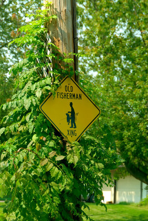 A sign on a post at the lakeside that says Old Fisherman Xing with a silhouette  image of a fisherman with fish on a pole.の写真素材