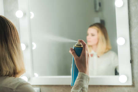Young woman styles her hair with hair spray looking in mirror