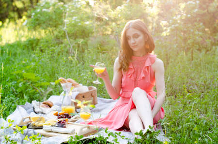 The red haired woman in park outside. Picnic setting on the grass with basket, bread, cheese, orange juice and fruitの写真素材