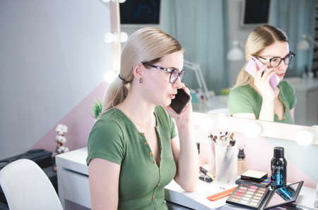 Blond female make up artist talks by phone with client. Professional stylist in her beauty visage...