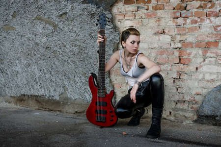 Punk girl dressed in black leather pants and white shirt, with red bass guitar at abadoned place