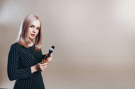 Beautiful blonde girl in black dress with make-up brushes on a beige background. Professional stylistの写真素材