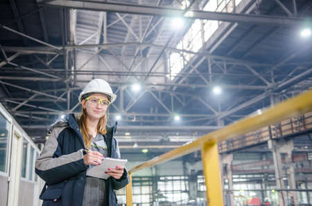 Young female technician in uniform, protective eyeglasses and helmet. Production workshop