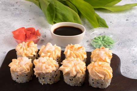 Rolls topped with cream sauce and fried shrimp. Sushi rolls set served on dark wooden board with wasabi, soy sauce and ginger. Gray background with green leafsの写真素材