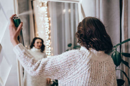 Young female applying hairspray on her hair while looking at mirror. Pretty young woman styles her hairの写真素材
