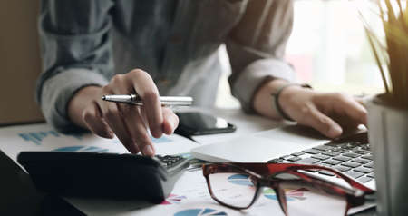 Close up of businessman or accountant hand holding pen working on calculator to calculate business data, accountancy document and laptop computer at office, business conceptの写真素材