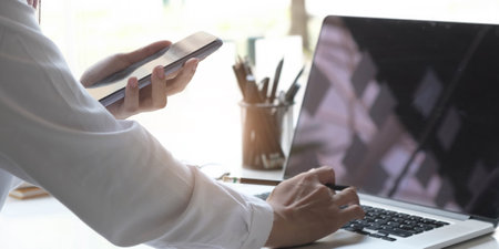 Close up of hand young female using mobile phone to searching information while she working with laptop on deskの写真素材