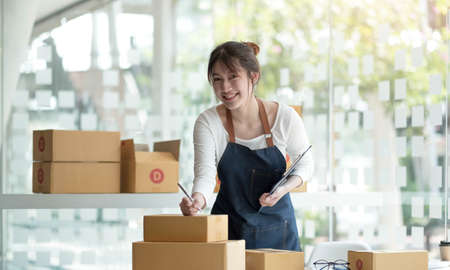 Smiling young Asian business owner woman prepare parcel box and standing check online orders of...