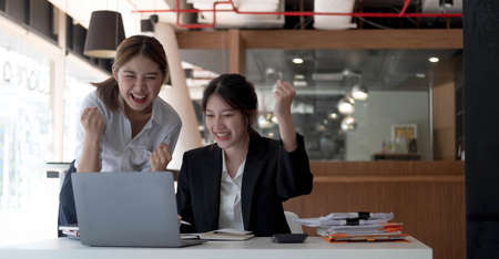 Two young Asian businesswomen show joyful expression of success at work smiling happily with a laptop computer in a modern office.の写真素材