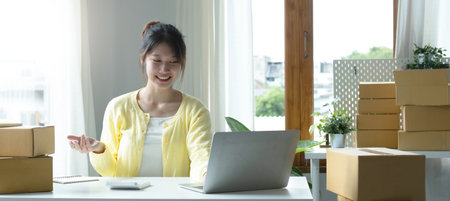 A portrait of Asian woman, e-commerce employee sitting in the office full of packages on the table using a laptop and smartphone, for SME business, e-commerce, technology and delivery business.の写真素材