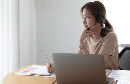 Asian businesswoman talking to colleague team in video call conference writing note on book with...