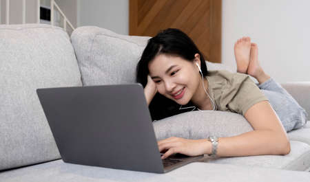 Asian woman lying on the sofa in the living room listening to music on a laptop device woman...
