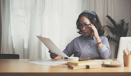 Seriously senior woman tworking at home, looking at laptop and waving paper correspondence, sitting at home, free space.の写真素材