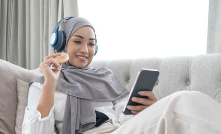 Attractive young Asian Muslim woman wearing hijab and headphones, watching movie on laptop computer on sofa.の写真素材