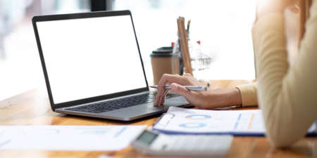 Mockup image of business woman using and typing on laptop with blank white screen and coffee cup on glass table in modern loft cafeの写真素材