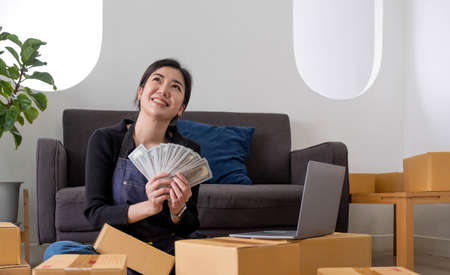 Young woman freelancer working and holding money with cardboard box  at home - SME business online...