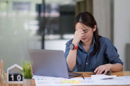Asian women sitting in a home office With stress and eye strain.Tired businesswoman holding eyeglasses and massaging nose bridge. There are tablets, laptops, and coffee.の写真素材