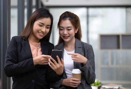 Two asian businesswoman in formal suit in office happy and cheerful during using smartphone and working in the office.の写真素材