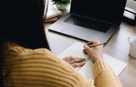 woman writing/taking notes while sitting in front her computer laptop at the wooden working table over living roomの写真素材