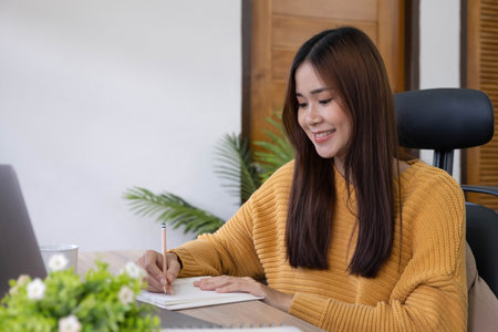 Beautiful woman writing/taking notes while sitting in front her computer laptop at the wooden working table over living room bookshelf as background.の写真素材