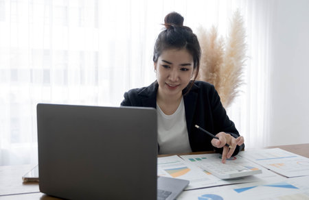 Beautiful Asian businesswoman analyzes charts using laptop calculator at the office.の写真素材