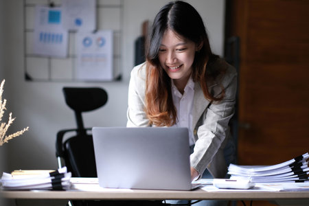 Beautiful Asian businesswoman standing and working on laptop with smiling face while working in...