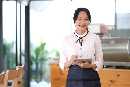 Beautiful Asian woman barista wearing an apron at the cafÃ© owner holding the tablet. Looking at camera.の写真素材