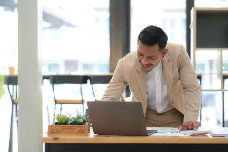 Portrait of a male business owner showing a happy smiling face as he has successfully invested his business using computers and financial budget documents at work.の写真素材