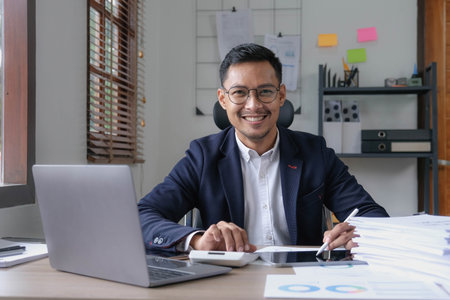 Portrait of a male business owner showing a happy smiling face as he has successfully invested his business using computers and financial budget documents at work.の写真素材