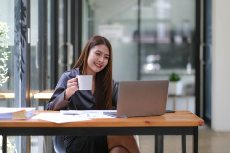 Happy excited successful Asian businesswoman triumphing with a laptop computer smartphone in the workplace officeの写真素材