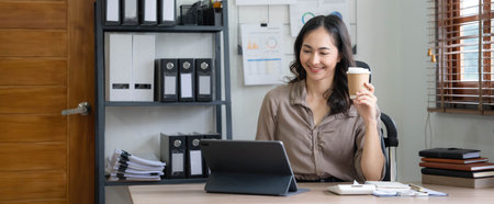 Beautiful young Asian businesswoman smiling holding a coffee mug and laptop working at the office.の写真素材