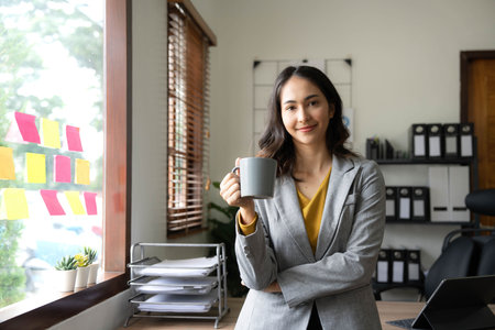 Beautiful young Asian businesswoman smiling holding a coffee mug and laptop working at the office.の写真素材
