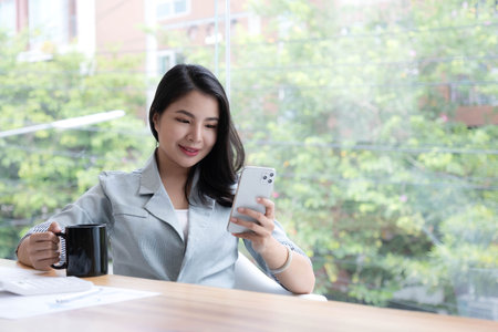 Beautiful young Asian businesswoman smiling holding a coffee mug and smartphone working at the office.の写真素材