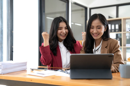 Two young Asian businesswomen show joyful expression of success at work smiling happily with a laptop computer in a modern office.の写真素材