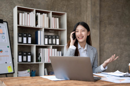 Smiling beautiful Asian businesswoman analyzing chart and graph showing changes on the market and holding smartphone at office.の写真素材