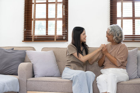 Portrait of Asian lovely family, young daughter hugging older mother. Attractive woman and senior elder mature mom sit on sofa, enjoy activity at home together in living room and smile, look at cameraの写真素材
