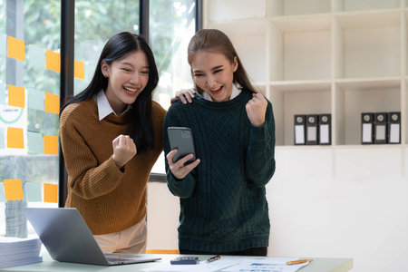 Two young Asian businesswomen show joyful expression of success at work smiling happily with a laptop computer in a modern office.の写真素材