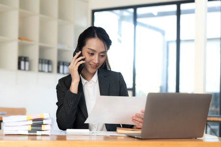 Smiling beautiful Asian businesswoman analyzing chart and graph showing changes on the market and holding smartphone at office.の写真素材