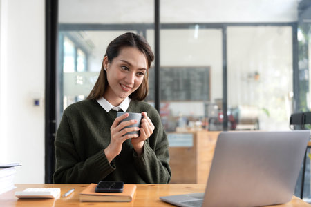 Beautiful young Asian businesswoman smiling holding a coffee mug and laptop working at the office.の写真素材