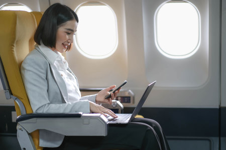 Asian woman passenger sitting in airplane near window and reading news from social networks or using travel applications in smartphoneの写真素材