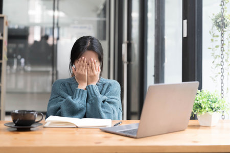 Young woman looks stressed, sitting at the desk, using a laptop, thinking and looking at the camera, feeling tired and bored with depression problemsの写真素材