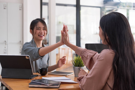 Two young Asian businesswomen show joyful expression of success at work smiling happily with a...