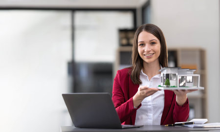 Young Asian real estate agent worker working with laptop at table in office and small house beside...
